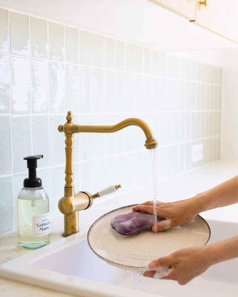 Des main de femme tiennent une assiette au dessus d'un évier, la main gauche tient l'assiette tandis que la main droite passe une éponge avec du liquide vaisselle dessus