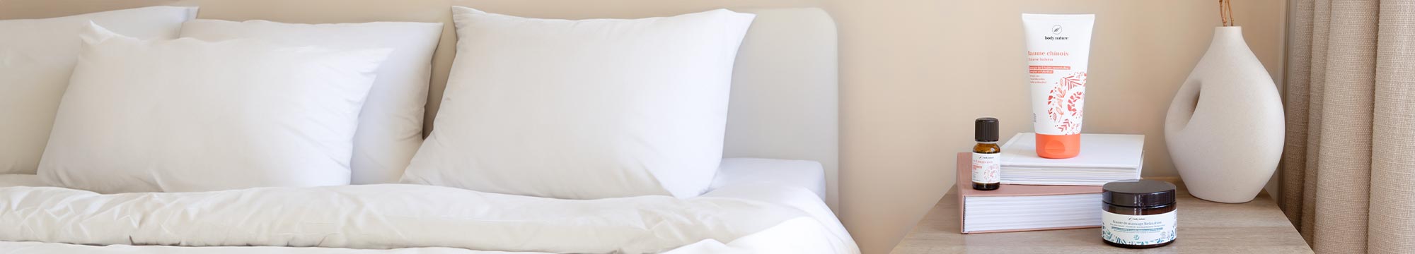 Un lit avec une table de nuit. Sur cette table de nuit, il y  3 produits Body Nature