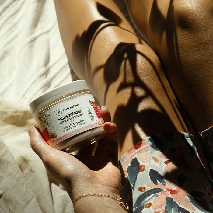 Pot du Baume hydratant pour le corps (200ml). Mise en scène : femme assise qui tient le pot dans sa main. Reflet de plantes sur l'image.