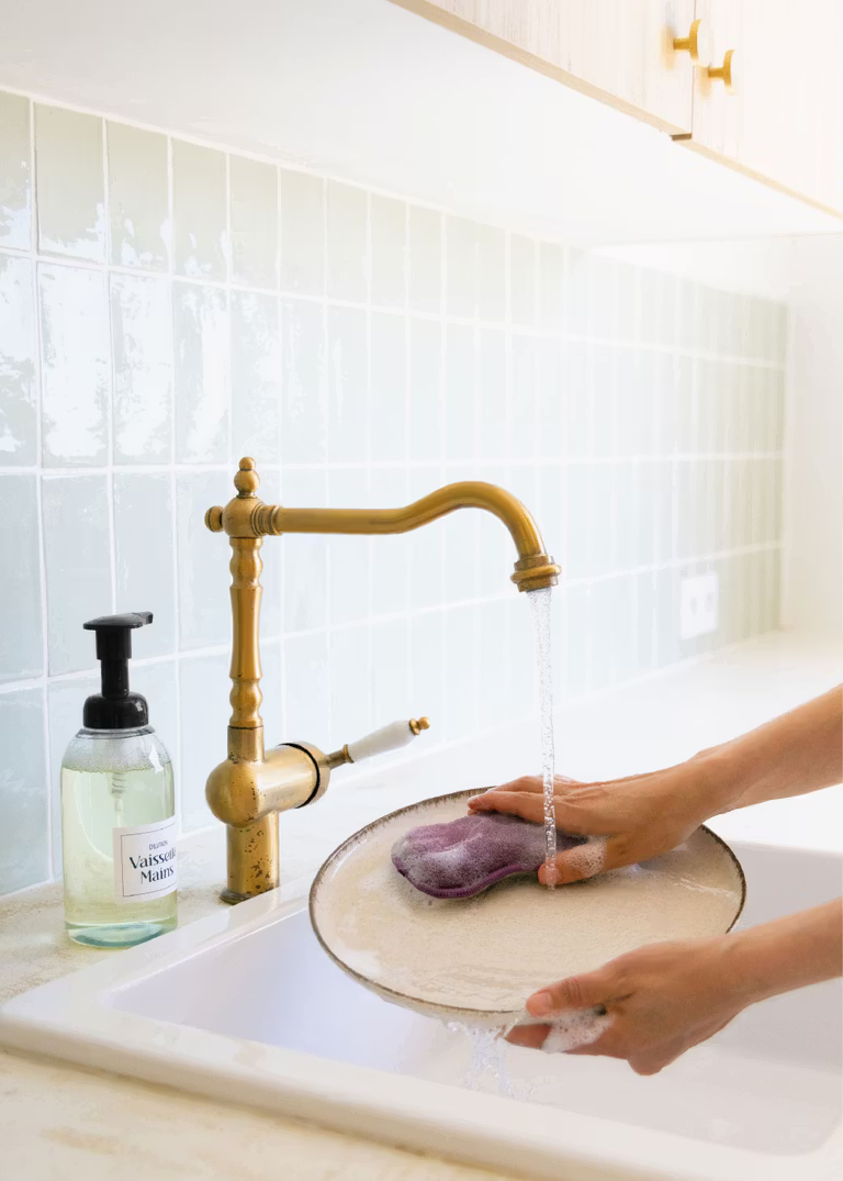Des main de femme tiennent une assiette au dessus d'un évier, la main gauche tient l'assiette tandis que la main droite passe une éponge avec du liquide vaisselle dessus